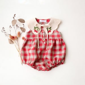 Può includere: Una tutina senza maniche con motivo a quadretti vichy rossi e beige, un ampio colletto color crema con motivi a fragola ricamati, un'abbottonatura frontale con tre bottoni in legno e un nastro color crema annodato sullo scollo.