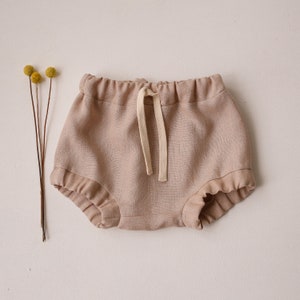 Puede incluir: Un par de bombachos de bebé de lino beige con cintura elástica fruncida y cordón blanco.