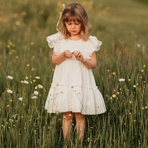 May include: A young girl wearing a white eyelet dress with a pink bow in her hair. She is standing in a field of wildflowers and holding a small flower.