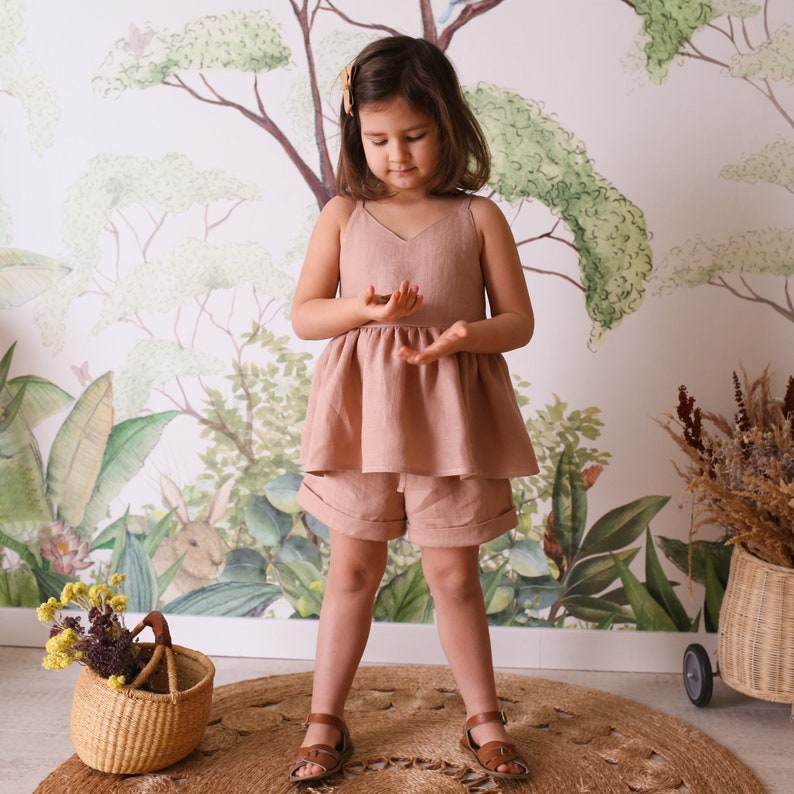 May include: A young girl wearing a light pink linen dress with a ruffled hem and matching linen shorts. She is standing on a brown rug in front of a wall with a painted jungle scene.