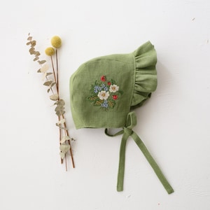 May include: A green linen baby bonnet with a floral embroidery design. The bonnet features a ruffled edge and ties. The embroidery includes white, red, and blue flowers. The bonnet is shown with dried flowers.