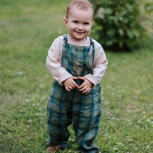 May include: A young child wearing a light pink long-sleeved shirt and green and blue plaid overalls. The overalls have a decorative embroidered design on the chest. The child is standing on green grass.