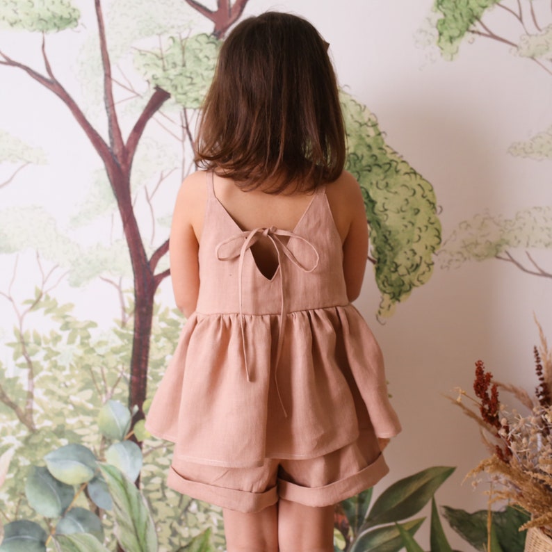 May include: A dusty pink linen top with a ruffled hem and a tie back. The top is worn with matching linen shorts.