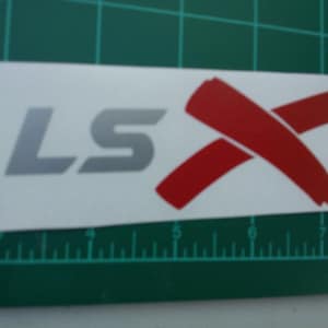 May include: A white sticker with the letters "LS" in silver and a red "X" design. The sticker is rectangular and placed on a green grid background. The sticker is approximately 6 inches long.