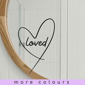 以下が含まれることがあります： 「loved」の文字が入った黒い線画のハートが鏡に表示されています。鏡は木製のフレームで、白いタイルの背景に配置されています。画像の下部には「more colours」というテキストがあります。