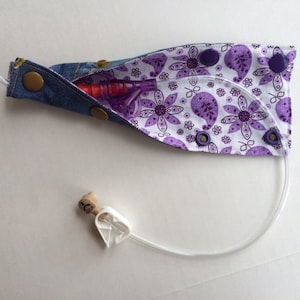 May include: A purple and white fabric pouch with a denim flap. The pouch has a clear plastic tube and a purple and red medical device inside. The pouch is secured with snaps.