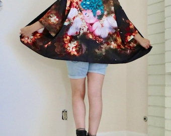 May include: A black kimono with a colorful floral print featuring a pink cherub with blue hair and wings. The kimono is worn over denim shorts and black ankle boots.