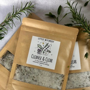 May include: Three brown paper pouches of bath tea with clear windows, labeled "CLEANSE & CLEAR" and "LITTLE WITCHERY." Each pouch contains a 7 oz. mixture. Sprigs of rosemary and other herbs are scattered around the pouches on a white cloth.