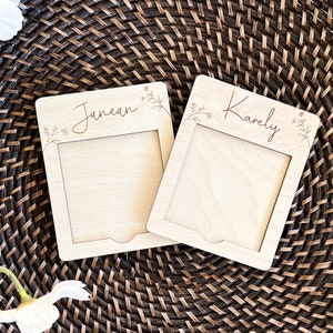 May include: Two wooden picture frames with a floral design and the names "Janea" and "Karely" engraved on them. The frames are sitting on a woven brown basket.