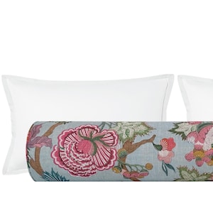 May include: A decorative bolster pillow with a floral pattern in shades of pink, red, blue, and green. The pillow is cylindrical and rests between two white rectangular pillows. The floral design is set against a light blue background.