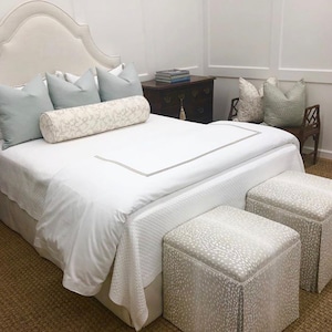 May include: A white bed with a white headboard and a white duvet cover. There are two light blue pillows and a white and gray patterned bolster pillow on the bed. Two white and gray patterned ottomans are at the foot of the bed.