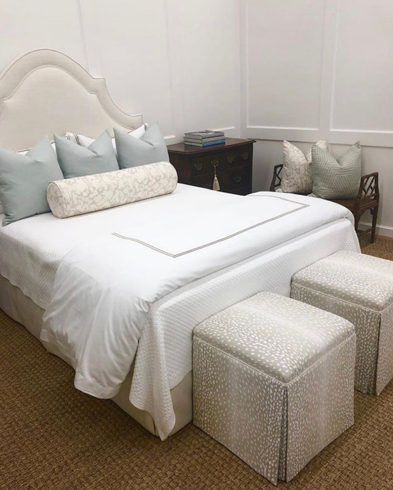 May include: A white bed with a white headboard and a white duvet cover. There are two light blue pillows and a white and gray patterned bolster pillow on the bed. Two white and gray patterned ottomans are at the foot of the bed.