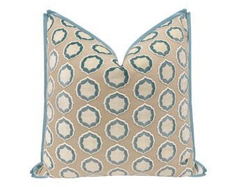May include: A square throw pillow featuring a beige base fabric with a repeating pattern of teal and white scalloped geometric shapes. The pillow is finished with a solid teal fabric piping along all four edges, creating a defined border.