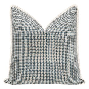 May include: A square throw pillow featuring a blue and cream houndstooth pattern. The pillow is finished with a light cream-colored fringe trim along all four edges, creating a textured border around the woven fabric design.