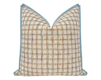 May include: A square throw pillow featuring a cream-colored fabric with a grid pattern of thin, multicolored lines in shades of blue, green, and brown. The pillow is finished with a solid light blue piping along all four edges.
