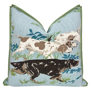 May include: Decorative throw pillow with a light blue background featuring a hunting dog design. The pillow has a green border and depicts three dogs in motion, with brown and white markings, amidst foliage.