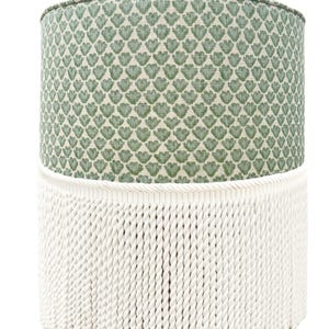Pode incluir: Um pufe cil&iacute;ndrico com um tampo com padr&atilde;o floral verde e uma base de franjas de corda branca. O padr&atilde;o verde &eacute; um desenho repetido de folhas estilizadas. Uma pe&ccedil;a de mobili&aacute;rio decorativa.