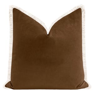 May include: A square, brown velvet pillow with a white fringe trim. The pillow has a soft, plush appearance and is designed for decorative use on a bed or sofa. The fringe adds a touch of elegance.