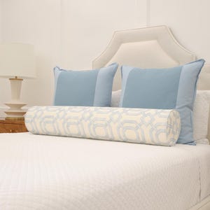 May include: A cylindrical bolster pillow featuring a cream-colored fabric with a light blue geometric lattice pattern, positioned in front of two solid light blue square pillows on a white quilted bedspread.