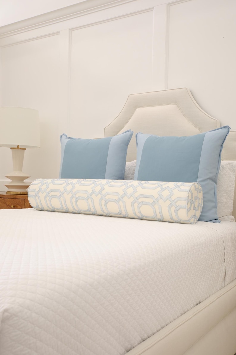 May include: A cylindrical bolster pillow featuring a cream-colored fabric with a light blue geometric lattice pattern, positioned in front of two solid light blue square pillows on a white quilted bedspread.