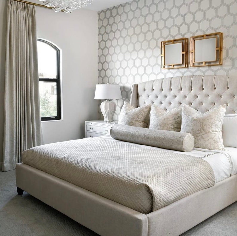 May include: A large, white upholstered bed with a tufted headboard and a decorative bolster pillow. The bed is in a bedroom with a patterned wallpaper and a white nightstand.