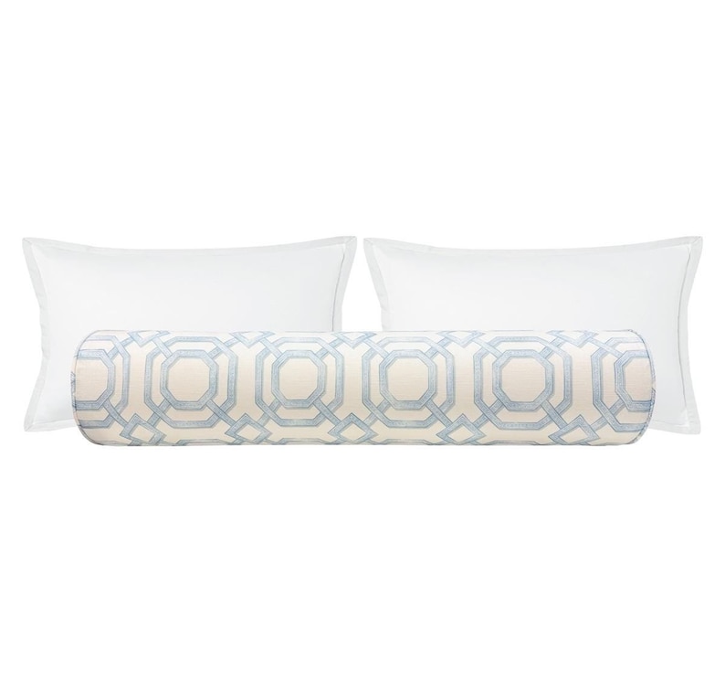May include: A cylindrical bolster pillow featuring a light blue geometric pattern on a cream background, positioned in front of two plain white rectangular pillows. The bolster has a repeating octagonal design across its entire length.