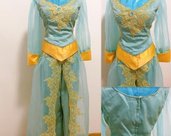 Può includere: Un costume turchese e oro con maniche lunghe e un'sovrapposizione trasparente. Il costume presenta ricami dorati intricati e una fascia gialla.
