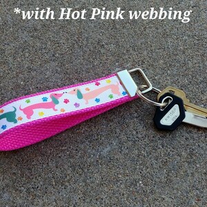 May include: A hot pink key fob with a white ribbon featuring dachshunds in pink and green, along with colorful paw prints. The key fob is attached to a silver metal ring with several keys. The text "with Hot Pink webbing" is visible.