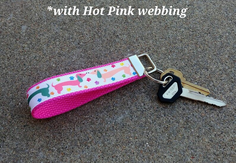 May include: A hot pink key fob with a white ribbon featuring dachshunds in pink and green, along with colorful paw prints. The key fob is attached to a silver metal ring with several keys. The text "with Hot Pink webbing" is visible.