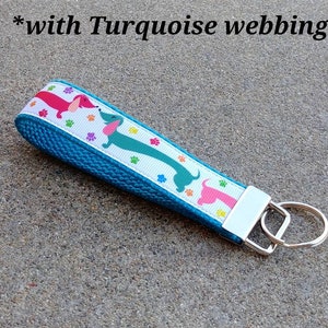 May include: A turquoise key fob with a white ribbon featuring colorful dachshunds and paw prints. The key fob has a silver-colored metal key ring and a matching metal end piece. The text "*with Turquoise webbing" is in the image.