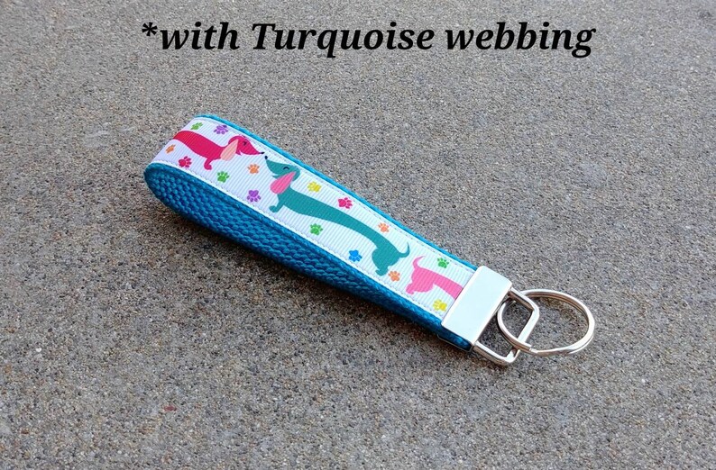 May include: A turquoise key fob with a white ribbon featuring colorful dachshunds and paw prints. The key fob has a silver-colored metal key ring and a matching metal end piece. The text "*with Turquoise webbing" is in the image.