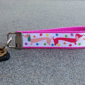 May include: A pink fabric keychain with a white ribbon featuring colorful dachshund dogs and paw prints. The keychain has a silver metal clasp and a set of keys attached.