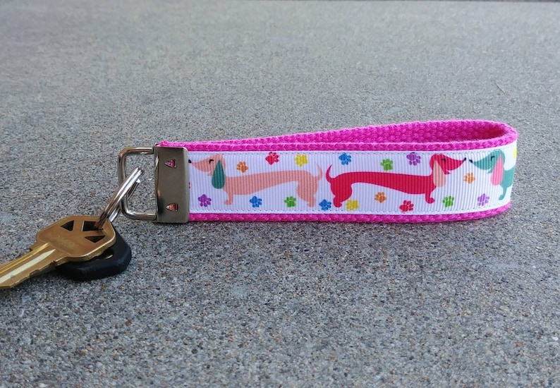 May include: A pink fabric keychain with a white ribbon featuring colorful dachshund dogs and paw prints. The keychain has a silver metal clasp and a set of keys attached.