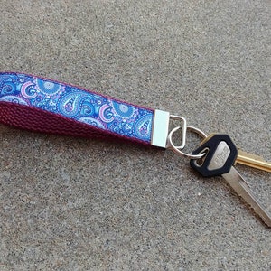 May include: A maroon key chain with a blue and pink paisley patterned ribbon. The key chain has a silver key ring with two keys attached.