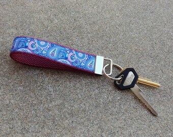 May include: A maroon key chain with a blue and pink paisley patterned ribbon. The key chain has a silver key ring with two keys attached.