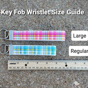 May include: Key fob wristlet size guide. Two wristlets with metal hardware, one pink and yellow plaid, the other blue and yellow plaid. A ruler shows the wristlets' length. Text reads "Large" and "Regular."