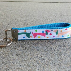 May include: A blue and white dachshund-themed keychain wristlet. The white ribbon features colorful dachshunds and paw prints. The wristlet has a blue woven strap and a silver key ring.
