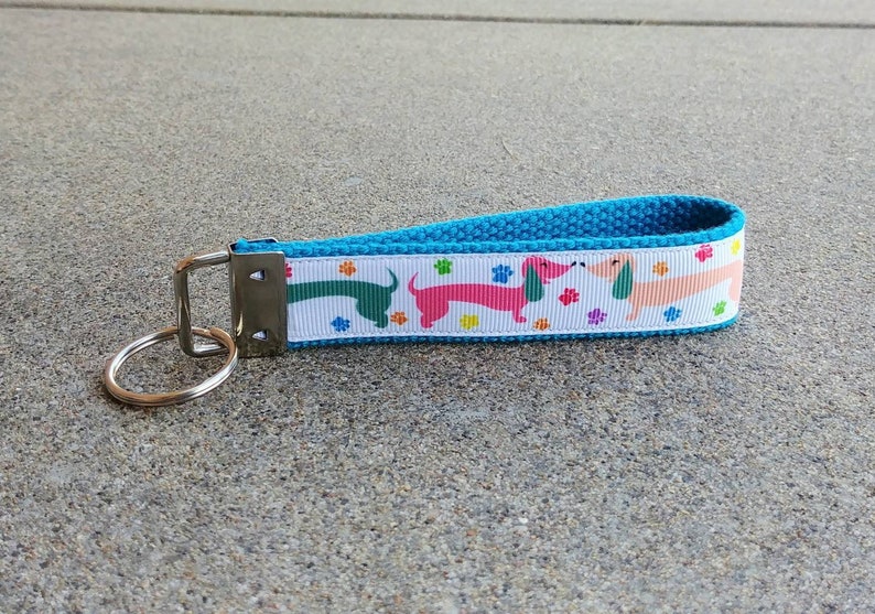 May include: A blue and white dachshund-themed keychain wristlet. The white ribbon features colorful dachshunds and paw prints. The wristlet has a blue woven strap and a silver key ring.