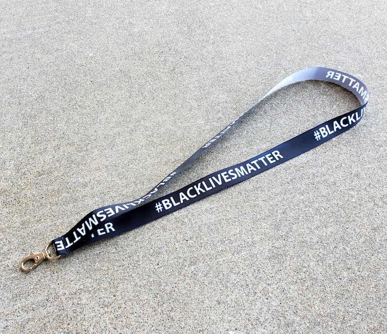 Puede incluir: Lanyard negro y blanco con el texto "#BlackLivesMatter" impreso en &eacute;l. El lanyard tiene un clip de metal dorado.