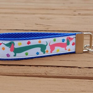 May include: A blue key fob with a white ribbon featuring colorful dachshunds and paw prints. The key fob has a silver-colored metal clasp and key ring. The background is a wooden surface.