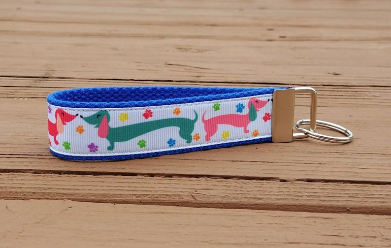 May include: A blue key fob with a white ribbon featuring colorful dachshunds and paw prints. The key fob has a silver-colored metal clasp and key ring. The background is a wooden surface.