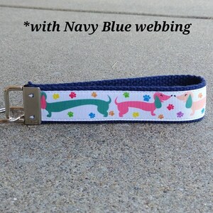 May include: A navy blue key fob with a white ribbon featuring colorful dachshunds and paw prints. The key fob has a silver-colored metal clasp and a key ring. The text "with Navy Blue webbing" is visible.