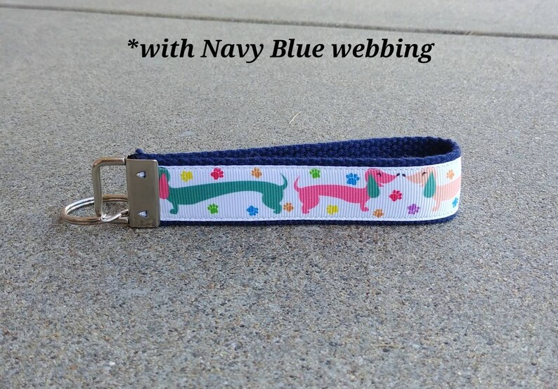 May include: A navy blue key fob with a white ribbon featuring colorful dachshunds and paw prints. The key fob has a silver-colored metal clasp and a key ring. The text "with Navy Blue webbing" is visible.