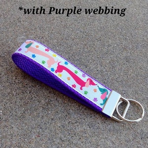 May include: A purple key fob with a white ribbon featuring pink, red, and blue dachshunds and paw prints. The key fob has a silver key ring and the text "*with Purple webbing".