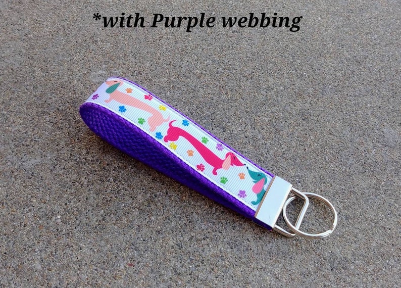 May include: A purple key fob with a white ribbon featuring pink, red, and blue dachshunds and paw prints. The key fob has a silver key ring and the text "*with Purple webbing".