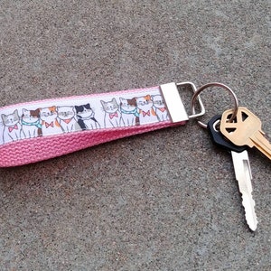 May include: Pink canvas keychain with a white ribbon featuring a line of cartoon cats wearing bow ties. The keychain has a silver key ring with two keys attached.