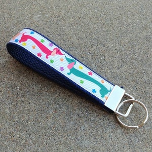 May include: A navy blue wristlet key fob with a white ribbon featuring colorful dachshunds and paw prints. The key fob has a silver-colored metal clasp and key ring.