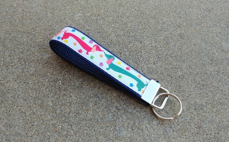 May include: A navy blue wristlet key fob with a white ribbon featuring colorful dachshunds and paw prints. The key fob has a silver-colored metal clasp and key ring.