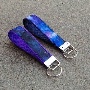 May include: Two galaxy print keychains with silver key rings. One keychain has a purple backing and the other has a navy blue backing.