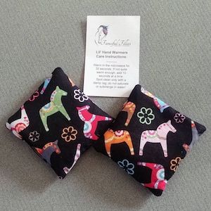 Puede incluir: Dos calientamanos de tela negra con un estampado de caballo escandinavo colorido. Los calientamanos est&aacute;n sobre una superficie gris con una tarjeta blanca que dice "Fanciful Fillies Lil' Hand Warmers Care Instructions: Warm in the microwave for 30 seconds. If not quite warm enough, add 10 seconds at a time Spot clean only with a damp rag, do not saturate or submerge in water!"